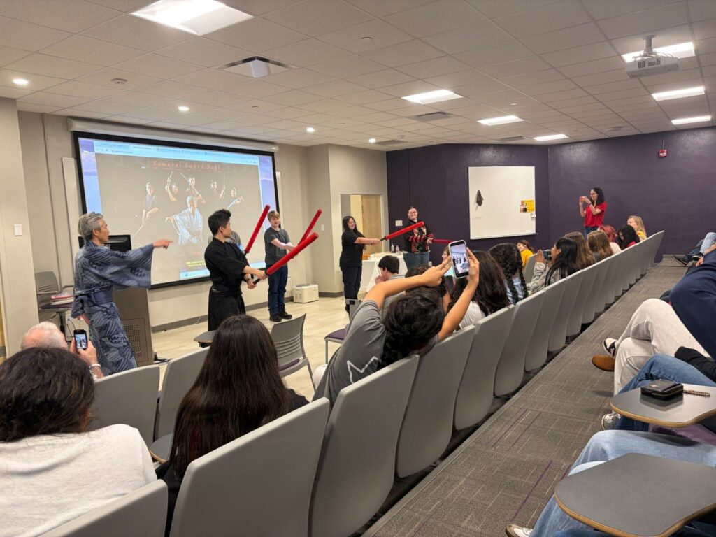 People in traditional attire and black uniforms demonstrate with foam swords at the front of a classroom, showcasing creative lesson plans as an audience watches and takes photos.
