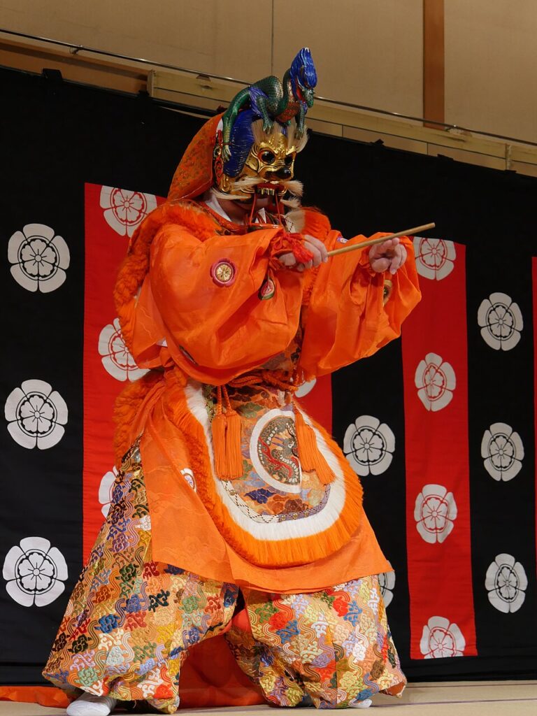 Performer in elaborate orange costume and ornate mask plays a flute on stage with a black and red background featuring white floral emblems, evoking the traditional elegance of Gagaku—Japanese Imperial Court Music—as preserved by the Imperial Household Agency.