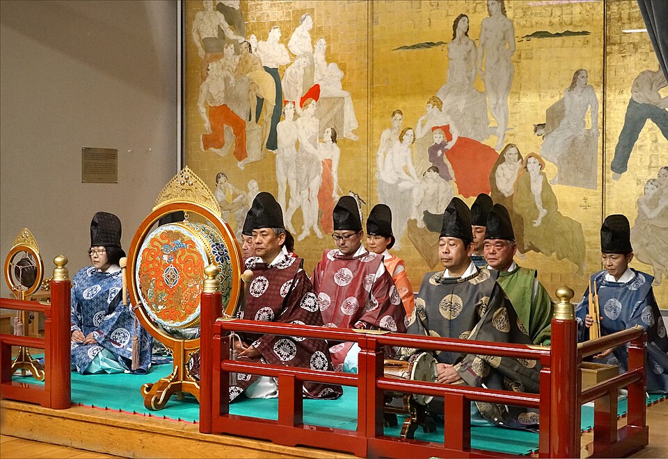 A group of people in traditional Japanese attire, part of a Gagaku Ensemble, sit behind drums and a red railing, with a gold mural depicting figures in the background.