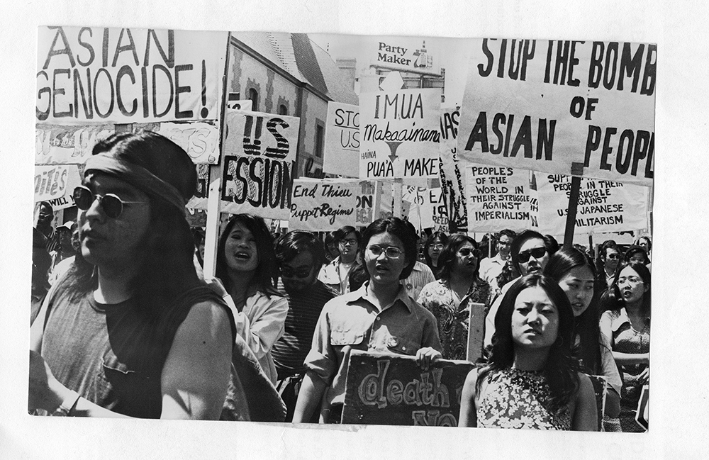 A group of protestors marches carrying signs with messages against Asian genocide, war, and militarism—capturing a powerful moment in Asian history. Black and white photo.