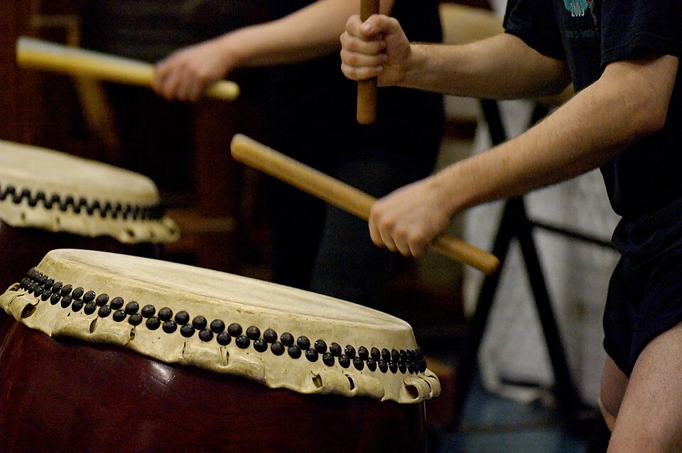 Two people engaged in Taiko Drumming are playing large traditional drums with wooden sticks, focusing on their arms and the drum surfaces during a powerful performance or practice reminiscent of cultural traditions from Japan.