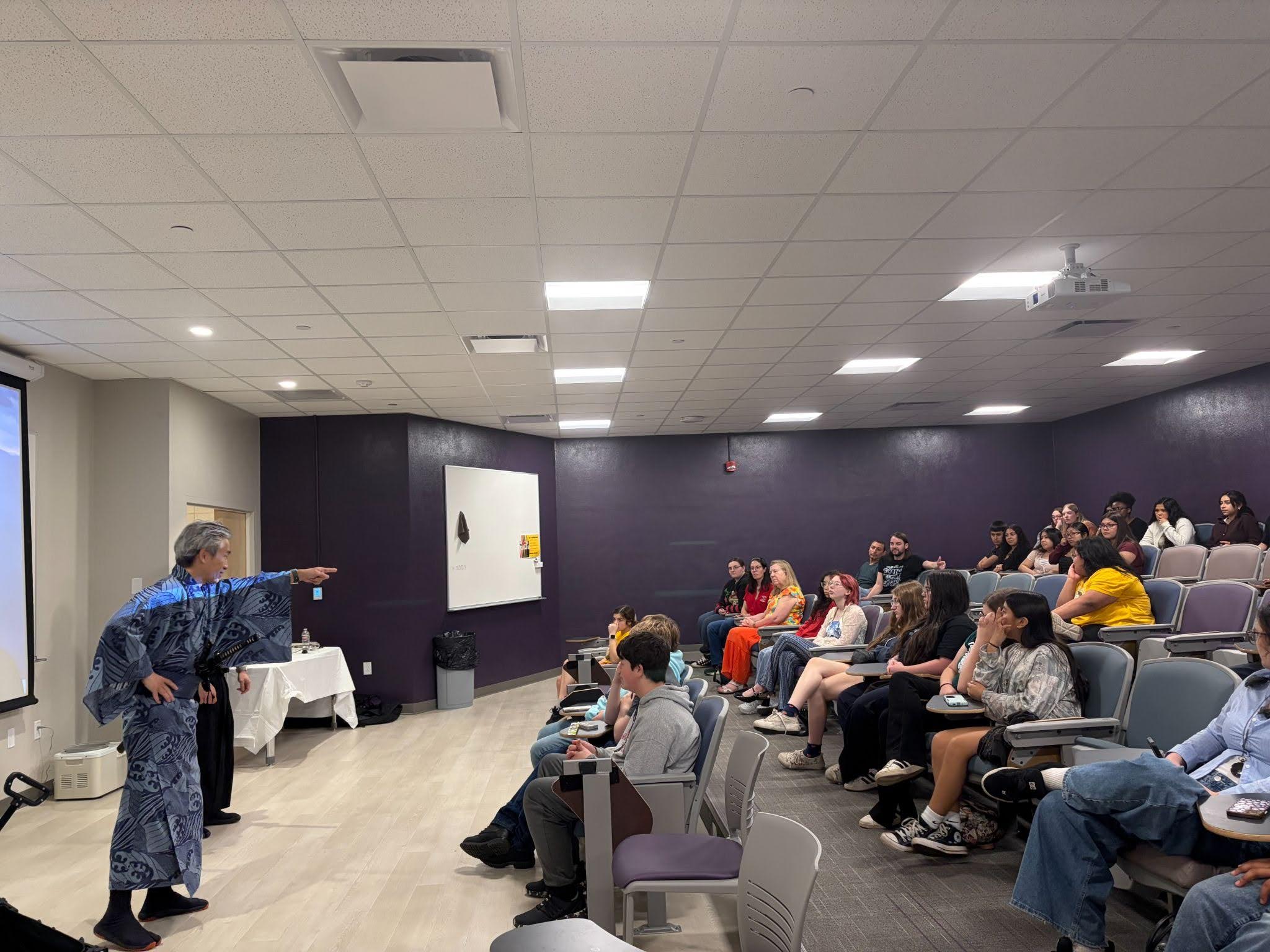 A person in a patterned robe stands at the front of a classroom, gesturing toward seated students who are watching attentively—an inspiring scene that reflects the spirit of teacher development supported by ARNCTA 2026 and opportunities like mini-grant applications.